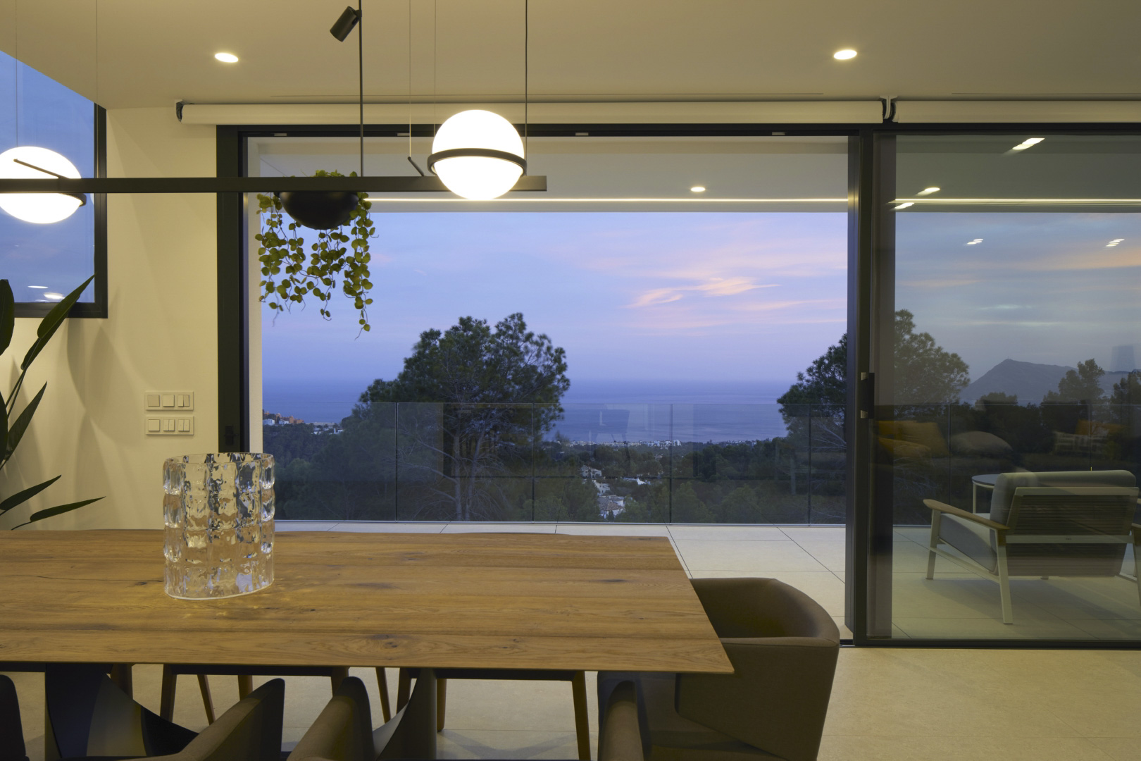 Dining space featuring a natural wood table and spherical pendant lighting, with floor-to-ceiling sliding doors opening to a terrace with sea views.