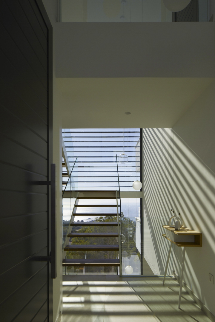 Entrance hall featuring a floating wooden staircase with glass railings. Natural light creates geometric shadows across the space, framing panoramic Mediterranean views.