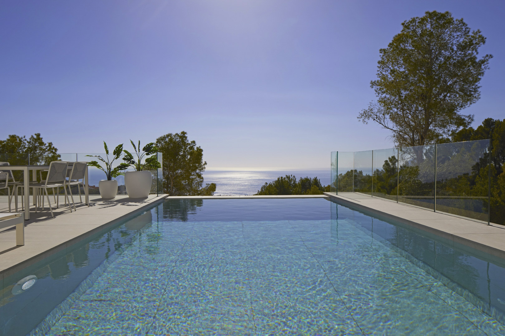 Infinity pool overlooking the Mediterranean Sea, featuring a sun terrace with modern dining furniture, glass balustrades, and lush pine surroundings.