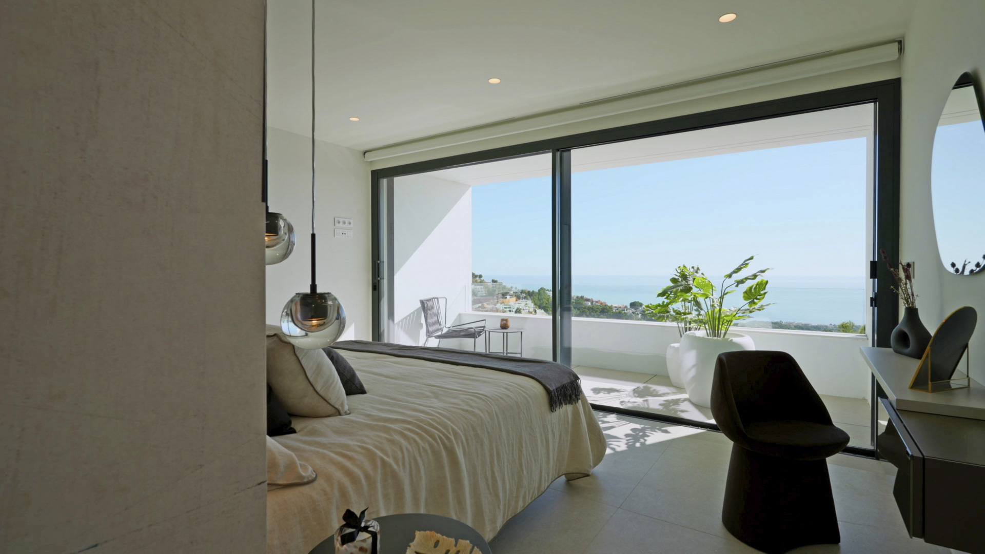 Master bedroom featuring floor-to-ceiling glass doors, a private terrace with sea views, and modern pendant lighting. The space includes a minimalist vanity.
