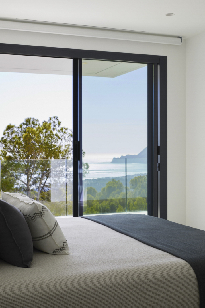 Master bedroom featuring floor-to-ceiling sliding glass doors, dark frames, and panoramic views of the Mediterranean Sea and coastal mountains.