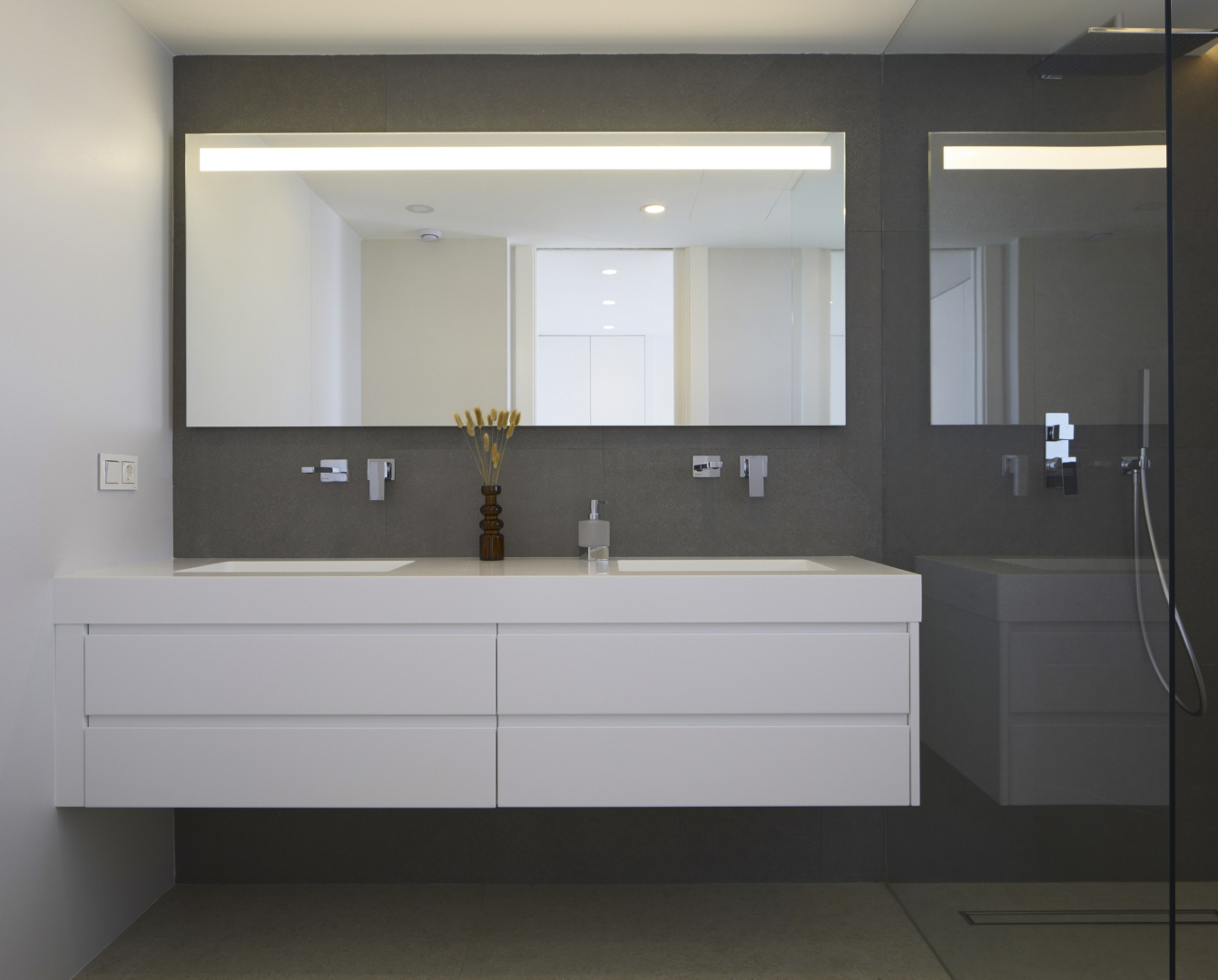Modern bathroom featuring a floating white double vanity, wall-mounted chrome fixtures, and a large backlit mirror against dark porcelain wall tiles.