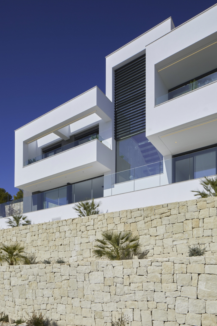 Modern facade featuring white geometric volumes and glass railings, elevated above a tiered natural dry stone wall with young palm trees.