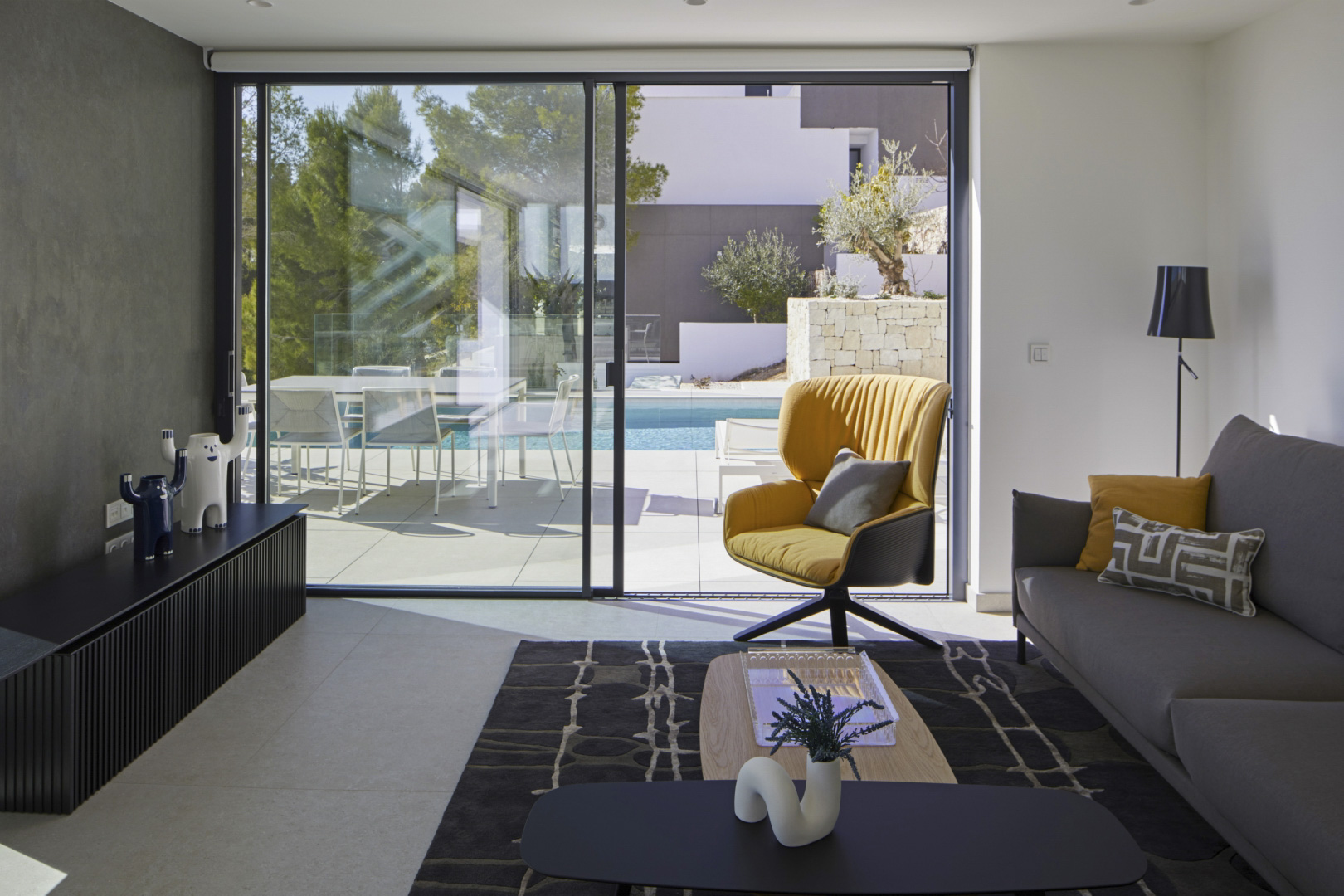 Modern living room featuring large sliding glass doors that open to the terrace and infinity pool. Includes designer furniture, a yellow accent chair, and tiled flooring.