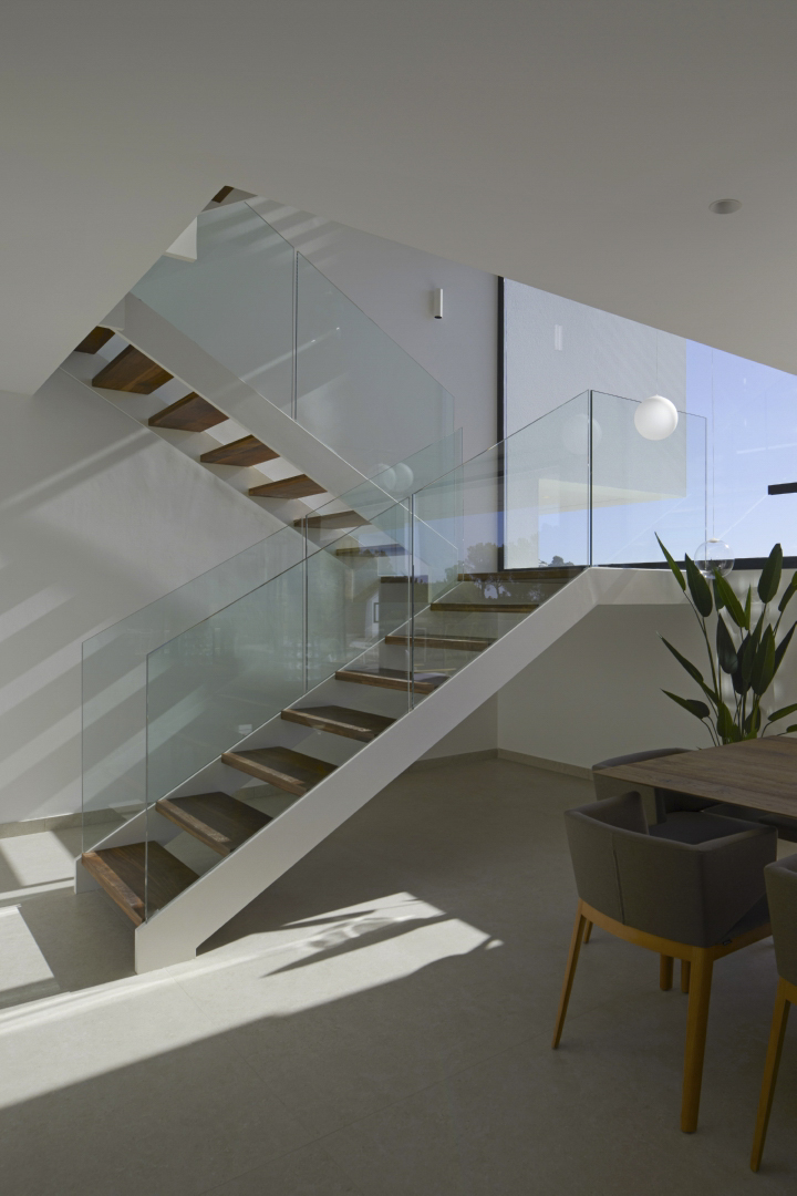 Modern staircase featuring dark wood treads and frameless glass railings, positioned in a bright interior with large windows and minimalist lighting.