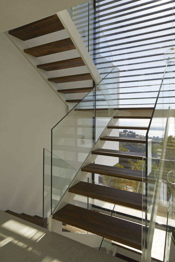 Modern staircase featuring dark wood treads and glass balustrades, positioned against a large window with coastal views and horizontal sun shades.