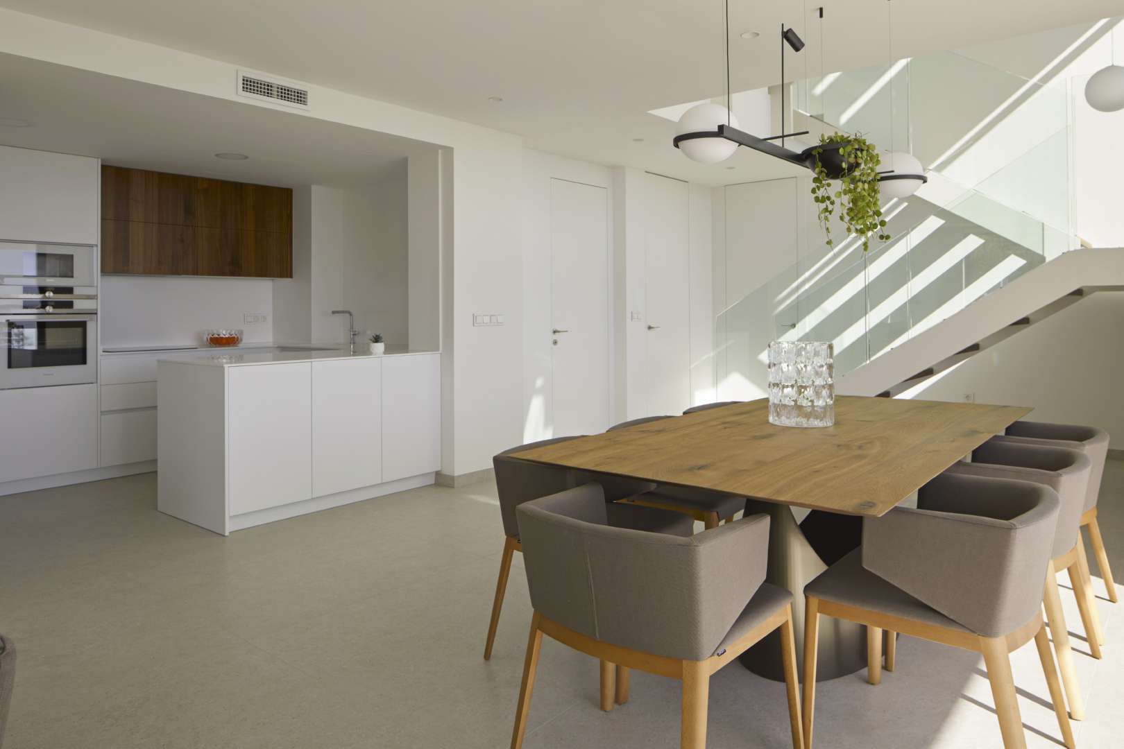 Open-plan dining area featuring a solid wood table, minimalist kitchen with white island, and a modern staircase with glass balustrade.