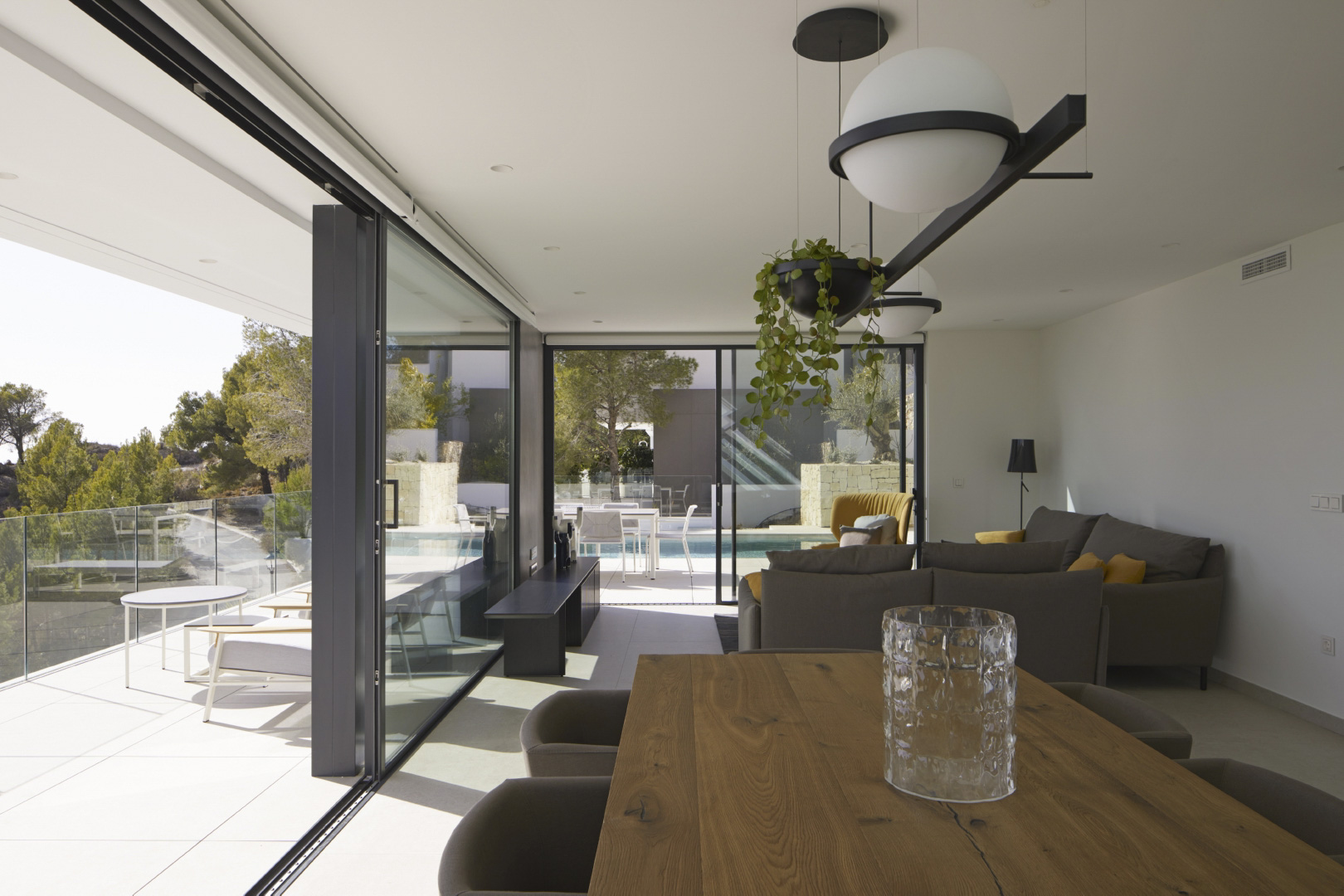 Open-plan living and dining area featuring a solid wood table, designer pendant lighting, and floor-to-ceiling glass doors opening onto the terrace and pool.