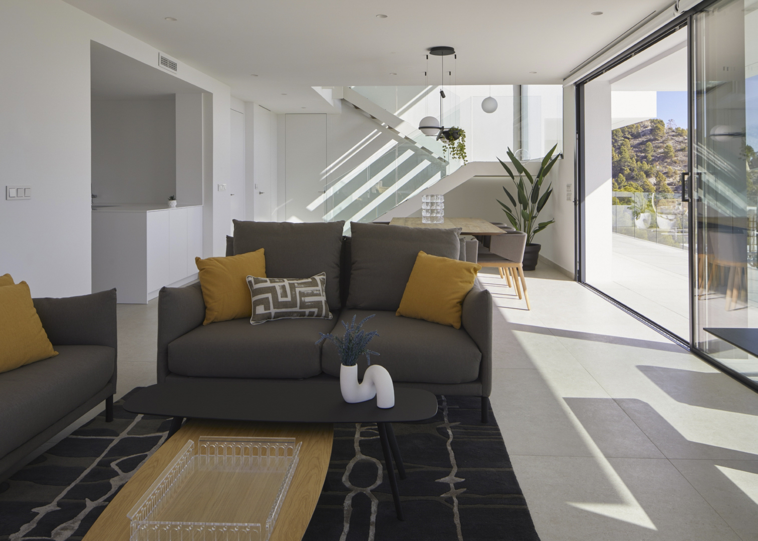 Open-plan living and dining area featuring grey sofas with mustard accents, a modern glass staircase, and large sliding doors to the terrace.