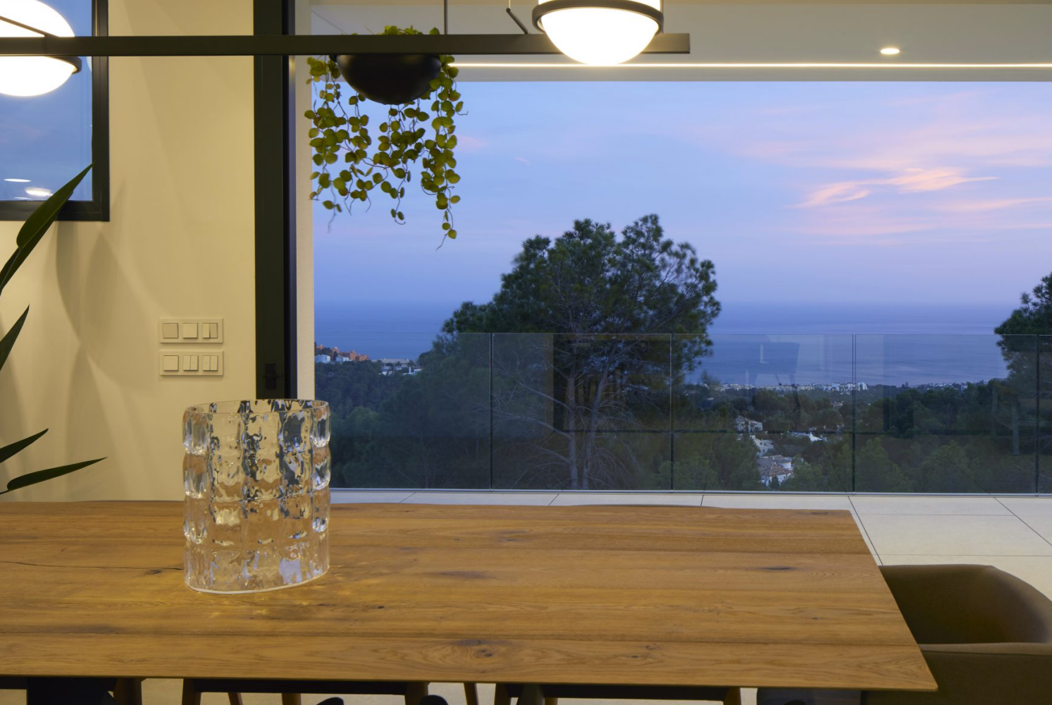 View from the dining area featuring a solid wood table and glass vase. Floor-to-ceiling windows open to a terrace with glass railings and sunset sea views.