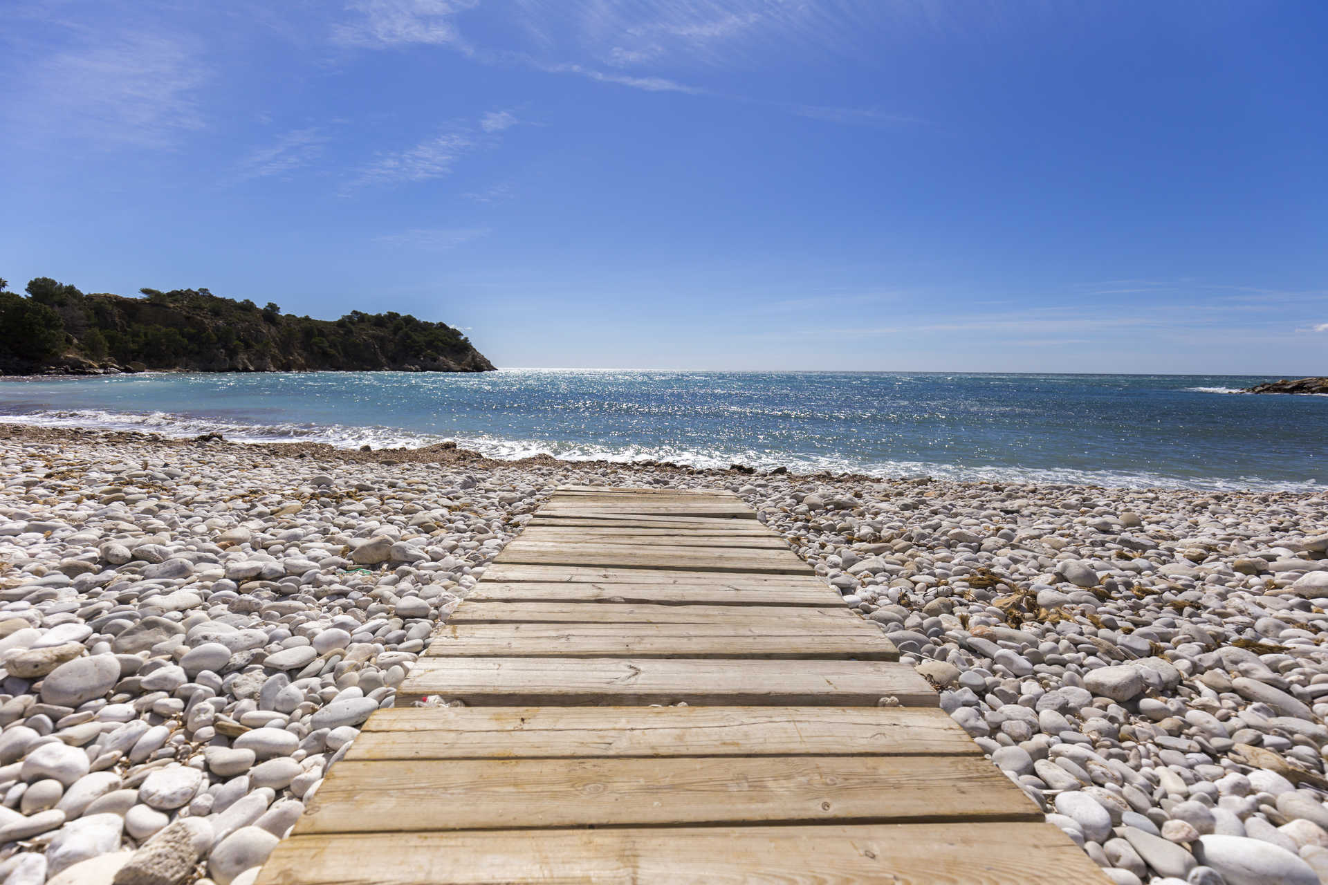 Wooden boardwalk crossing a white pebble beach towards the turquoise Mediterranean waters and the scenic coastline of Altea.