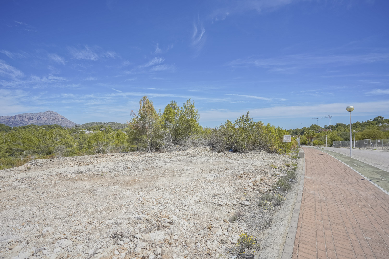 Elevated 1,000 m2 building plot in Jávea, featuring cleared terrain with views of the Montgó mountain, adjacent to a paved sidewalk and residential infrastructure.