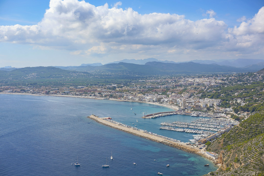 Panoramic aerial view of Jávea port and bay, featuring the marina, Mediterranean coastline, and distant mountain ranges under a bright sky.