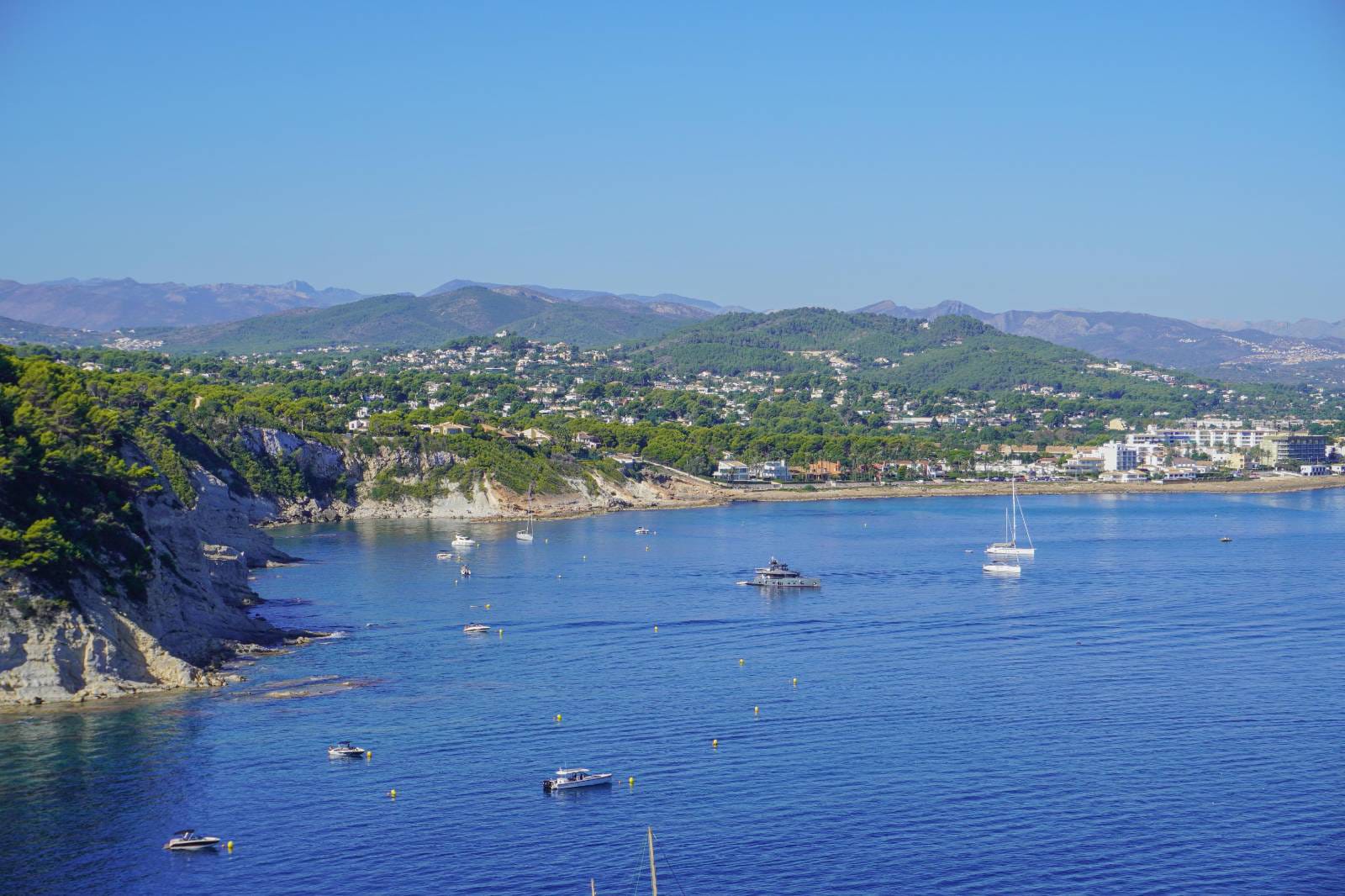 Panoramic coastal view of the Jávea bay, featuring rugged cliffs, anchored yachts, and a distant mountain horizon.