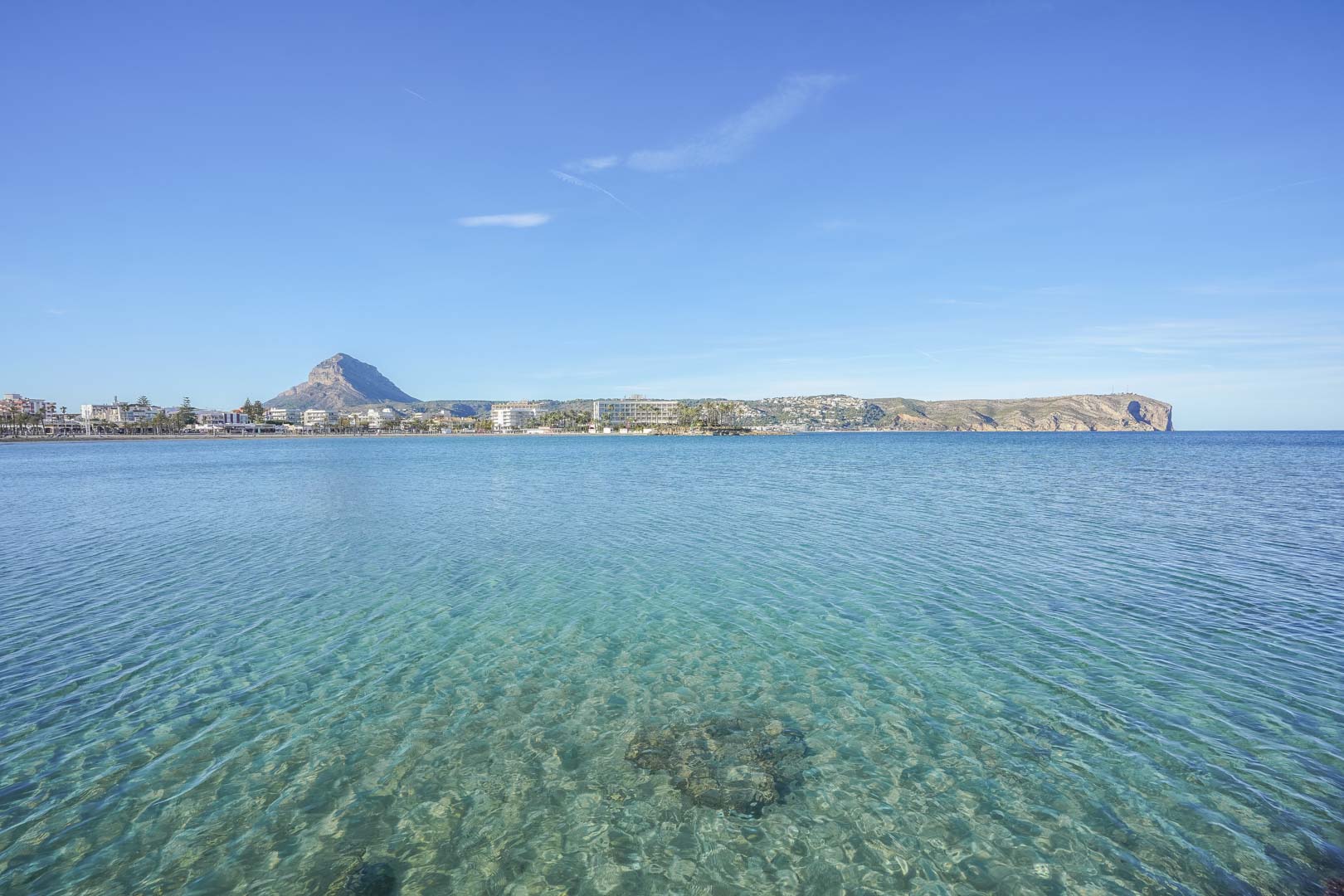 Panoramic view of Jávea's bay featuring crystal-clear turquoise waters, the Montgó mountain, and the coastal skyline under a bright blue sky.