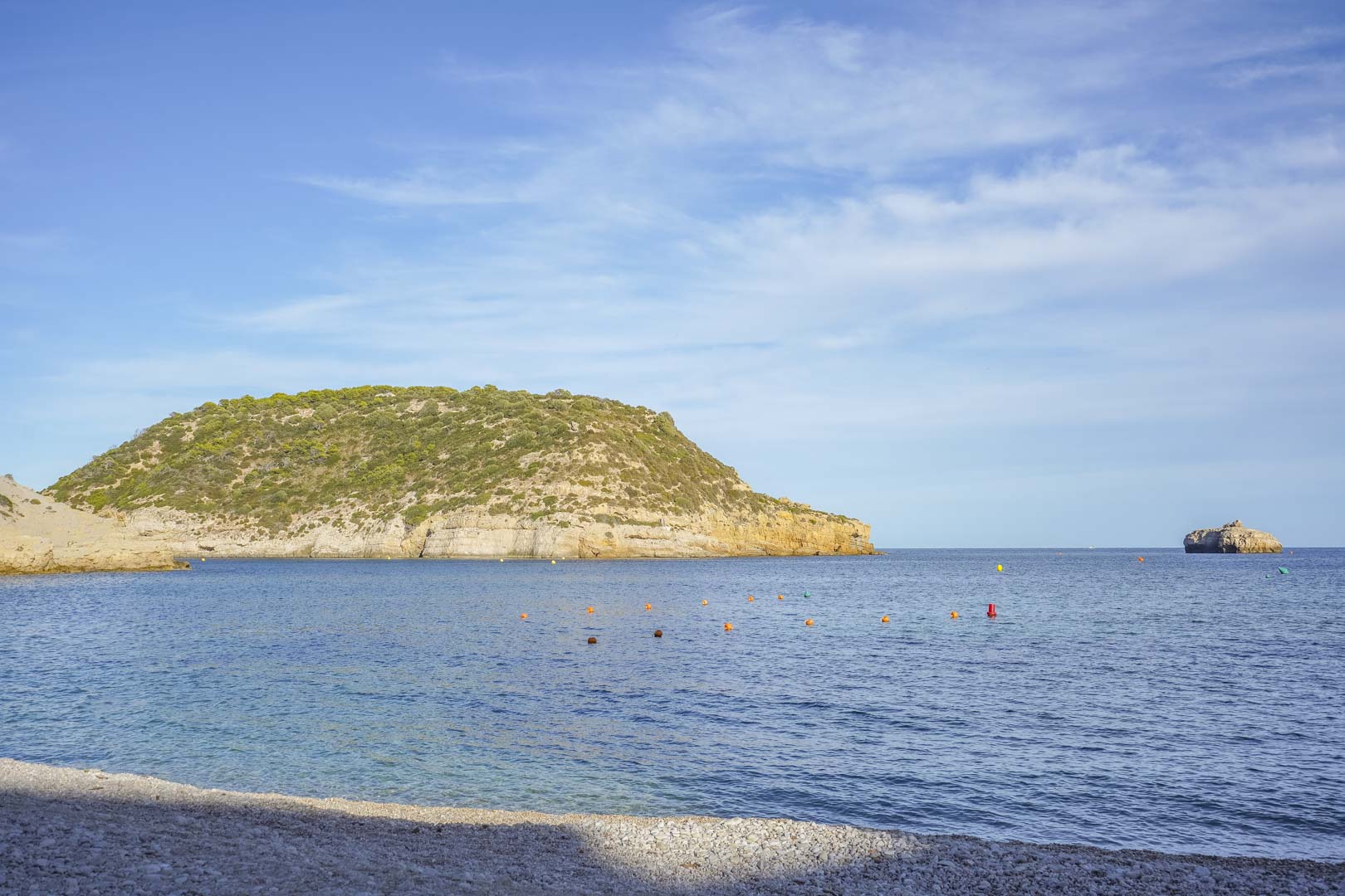 Panoramic view of a pebble beach in Jávea, featuring crystal-clear Mediterranean waters and rugged coastal cliffs under a bright blue sky.