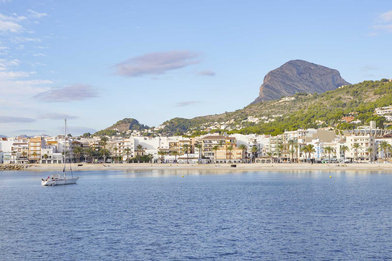 Panoramic view of Jávea's bay featuring the iconic Montgó mountain and a sailboat on calm Mediterranean waters near the coastal promenade.
