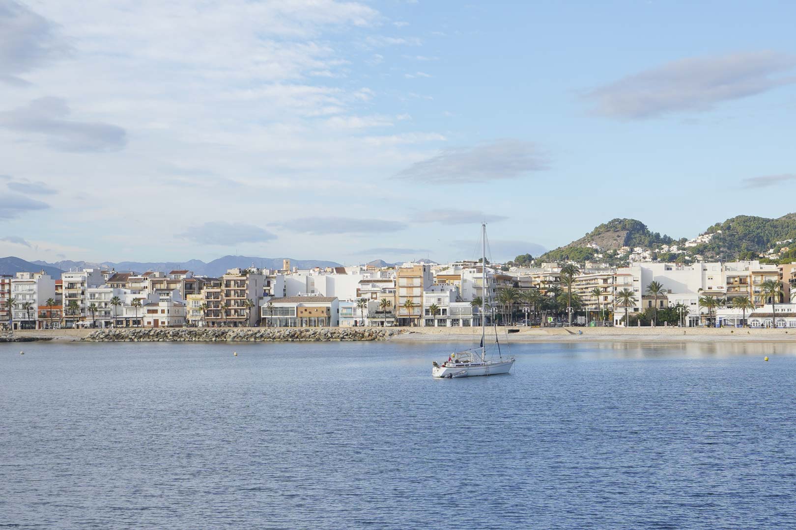 Panoramic view of the Jávea coastline featuring a sailboat at anchor, waterfront Mediterranean buildings, and the scenic mountain backdrop.