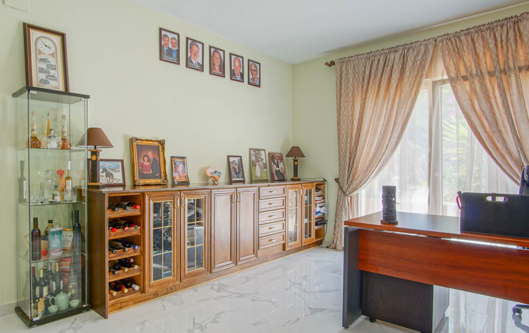 Bright home office featuring marble-effect flooring, a wooden desk, and a large sideboard with integrated wine storage and display cabinets.