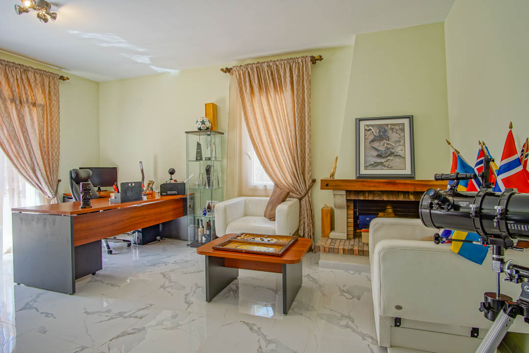 Bright home office featuring marble-effect tiling, a large wooden desk, a traditional fireplace, and a seating area with white leather armchairs.