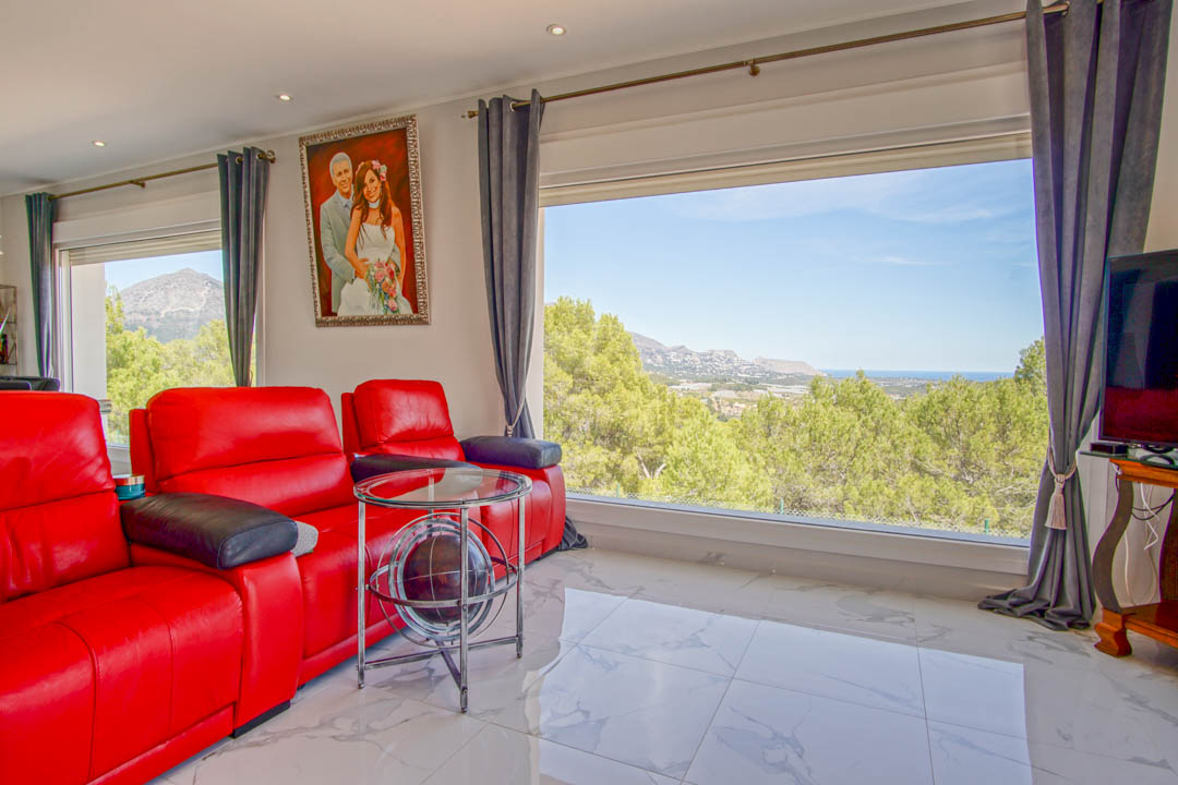 Bright living area featuring red leather recliners, marble-effect flooring, and large windows offering panoramic views of the Mediterranean sea and mountains.