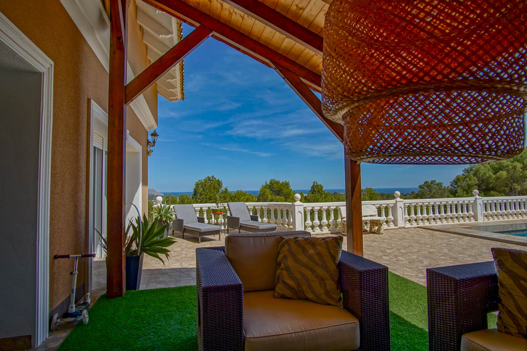 Covered terrace featuring exposed wooden beams, wicker seating, and panoramic Mediterranean Sea views beyond a classic white balustrade.