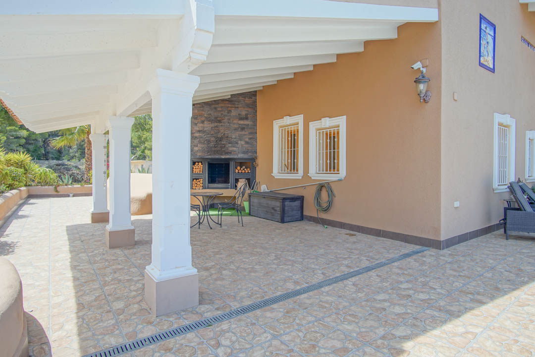 Covered terrace featuring white wooden beams, classic columns, and a built-in stone fireplace with dedicated wood storage and tiled flooring.