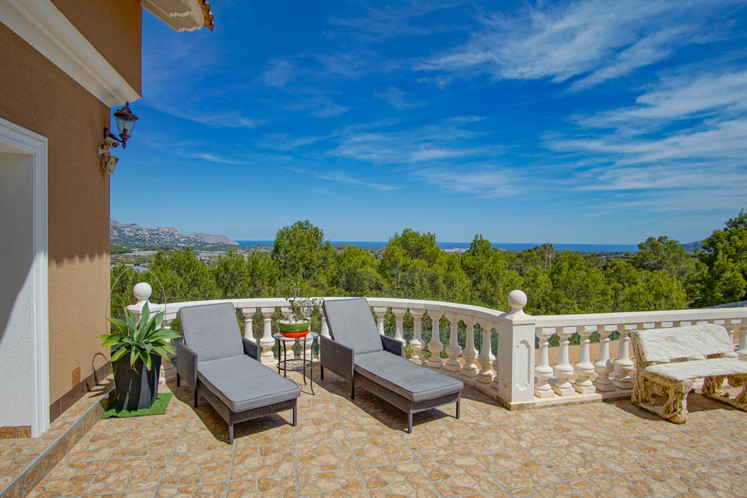 Elevated terrace featuring irregular stone tiling, a white balustrade, and grey sun loungers overlooking a dense pine forest and the Mediterranean Sea.