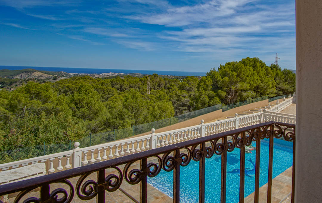 Elevated view from a balcony with a wrought iron railing, overlooking the pool area and lush pine forests extending toward the Mediterranean Sea.