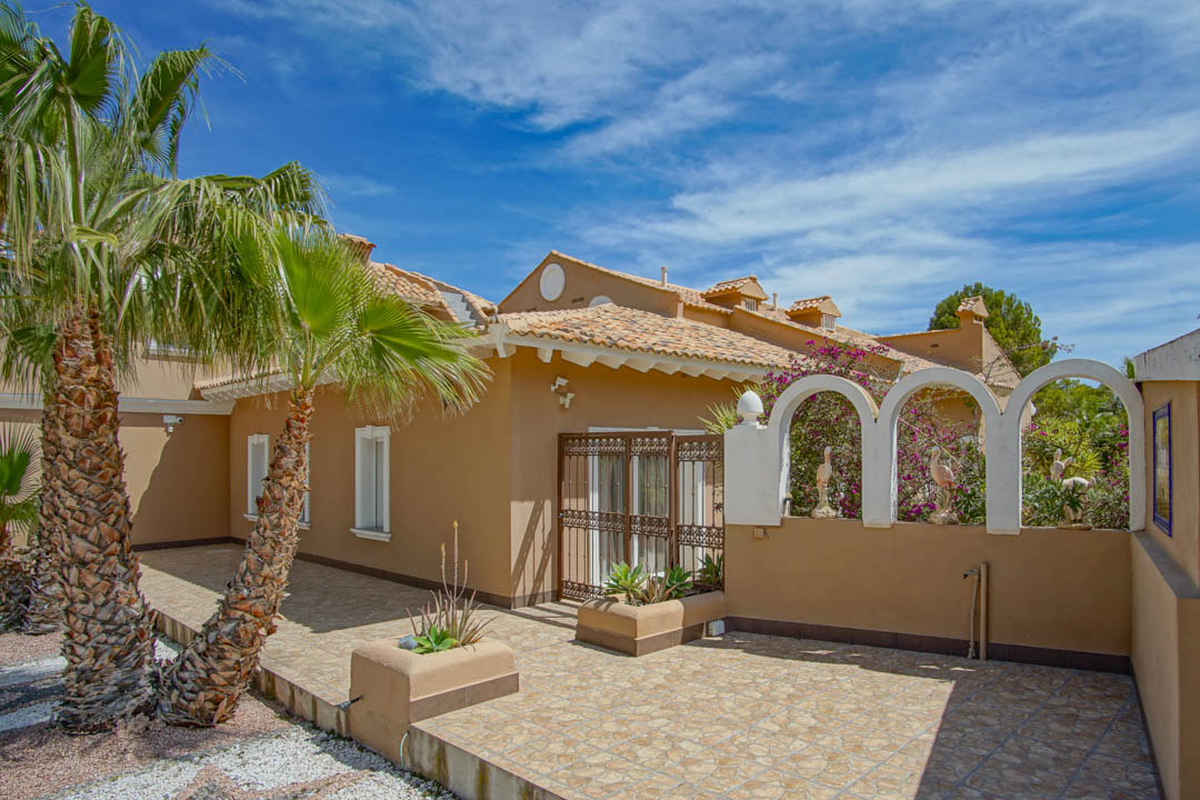 Exterior courtyard with ceramic tiling and ochre walls, featuring white decorative arches with ornamental figures, palm trees, and a traditional wrought iron gate.
