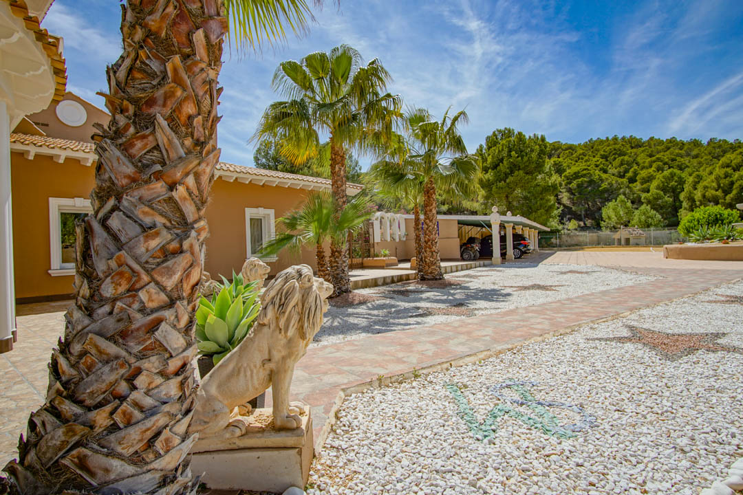 Exterior entrance featuring stone lion statues, palm trees, and a decorative gravel driveway with mosaics leading to a covered carport adjacent to a pine forest.
