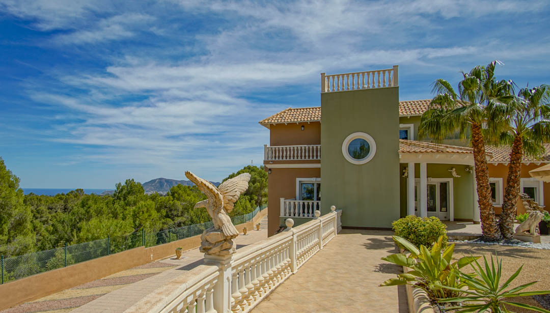 Exterior facade featuring classic balustrades, an ornamental eagle sculpture, and a palm-lined entrance overlooking the Mediterranean pine forest and sea.