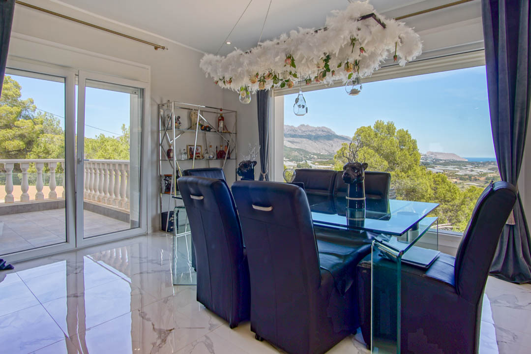 Formal dining room featuring marble flooring, a glass table, and black leather chairs, with expansive windows showcasing panoramic mountain and sea views.