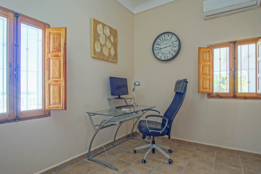 Functional office space featuring a glass-top desk, traditional wooden shutters on dual windows, and rustic tiled flooring.