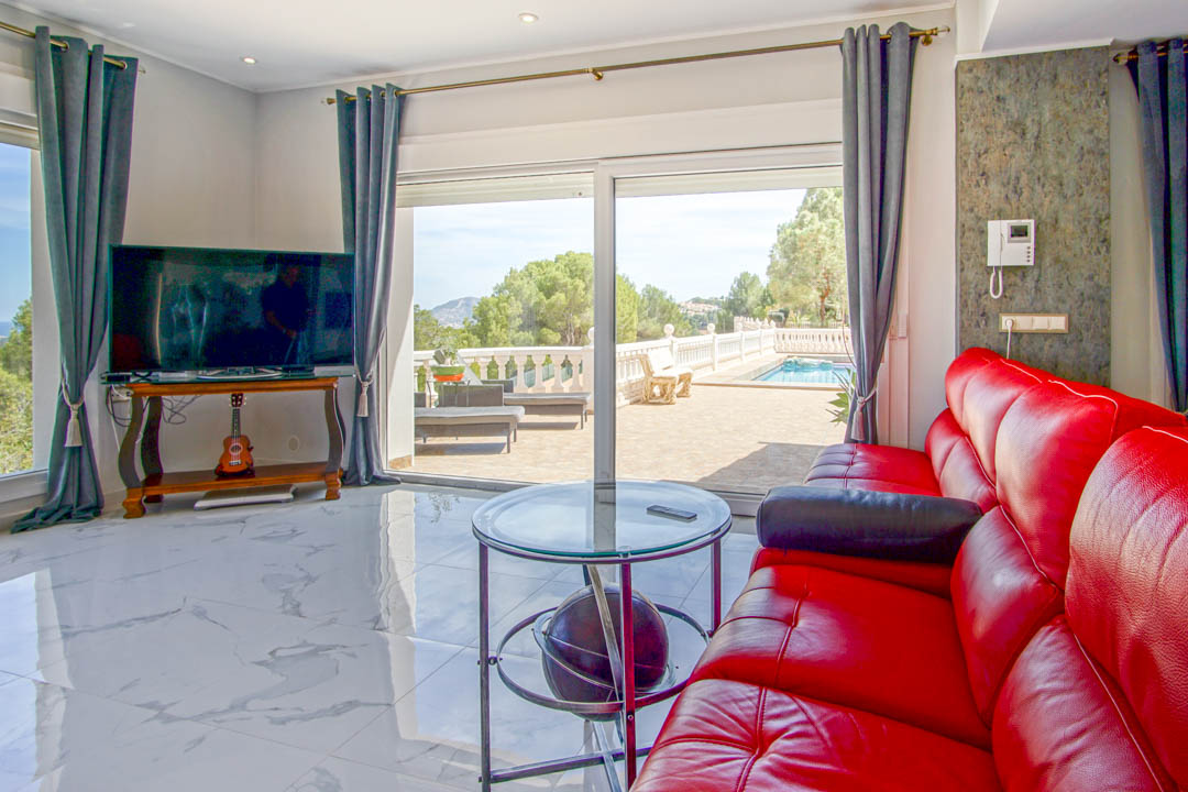 Living area featuring white marble-effect flooring, a red leather sofa, and large sliding doors opening onto a sun terrace with a private pool.