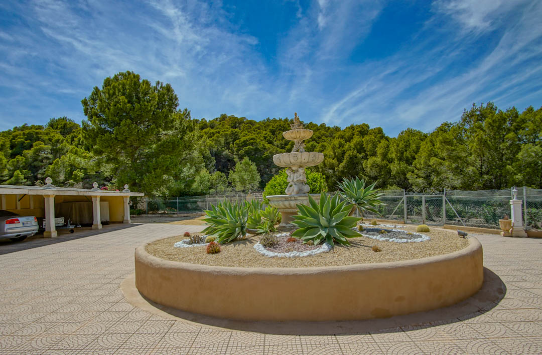 Main entrance featuring a three-tier classical fountain and circular planter with agaves, adjacent to a covered carport and pine forest.