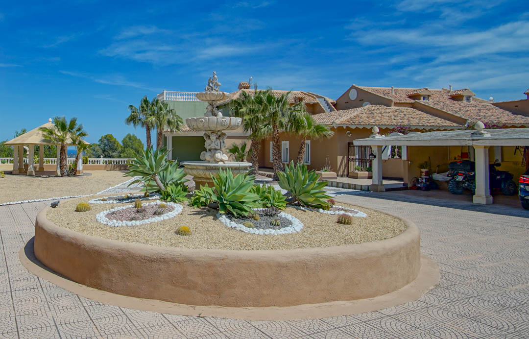Main entrance featuring an ornate stone fountain, a circular planter with agaves and cacti, a covered carport, and a garden pavilion under a clear sky.