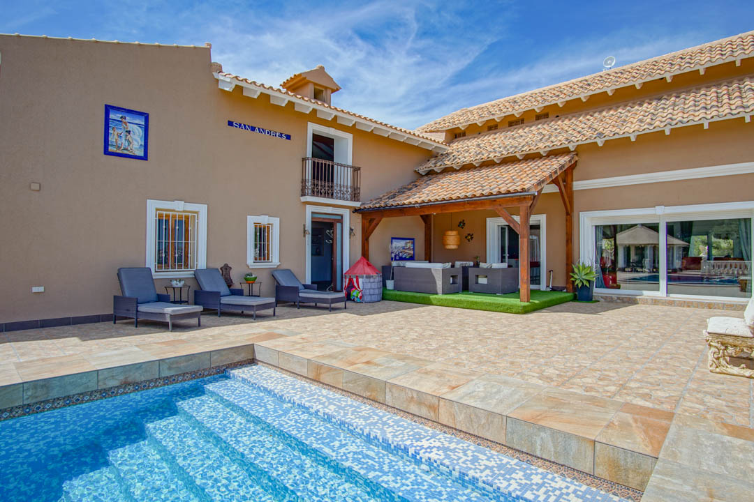 Main terrace featuring a blue mosaic pool, sun loungers, and a covered porch with exposed wooden beams and a lounge area on artificial turf.