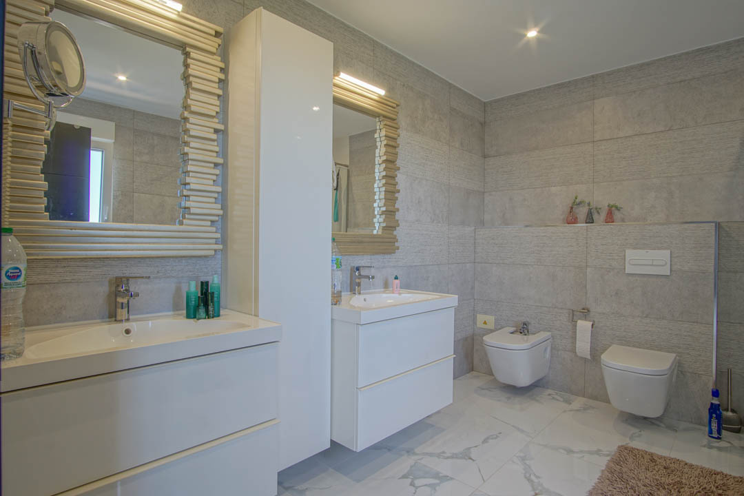 Modern bathroom featuring a double white vanity, mirrors with decorative slatted frames, and grey textured wall tiles. Includes a wall-mounted toilet and bidet.
