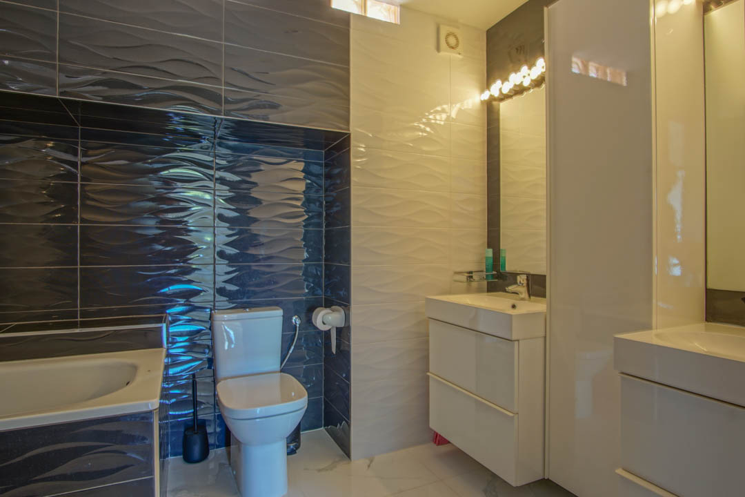 Modern bathroom featuring textured wave-patterned tiles in white and dark grey, a sleek white vanity unit, and integrated vanity lighting above the mirror.