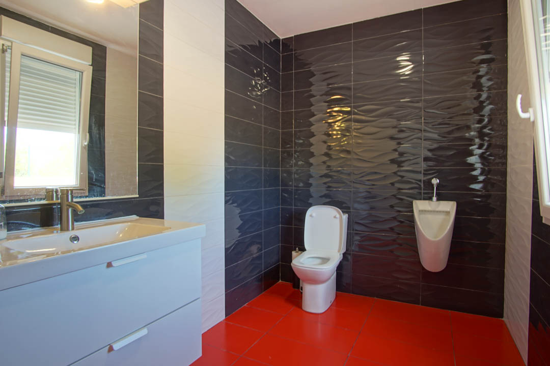 Modern bathroom featuring vibrant red floor tiles, dark wavy-textured wall ceramics, a white vanity unit, and a wall-mounted urinal.