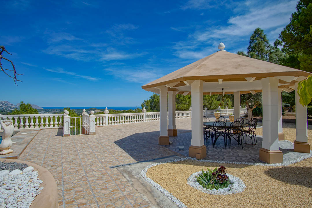 Outdoor pavilion featuring white pillars and wrought iron dining furniture, set on a tiled terrace with expansive views of the Mediterranean coastline.