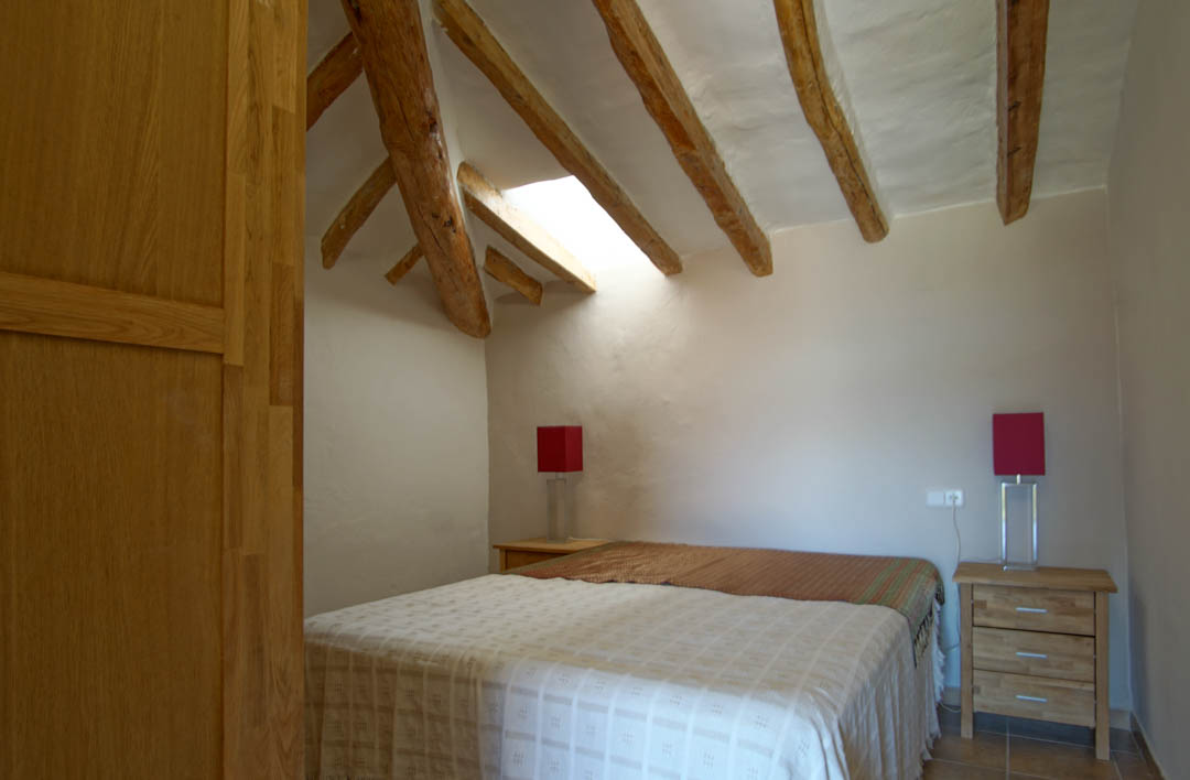 Rustic bedroom featuring exposed timber beams, a skylight for natural illumination, and textured white walls. Furnished with wooden nightstands and red bedside lamps.