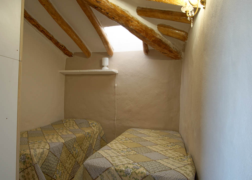 Rustic bedroom featuring sloped ceilings with exposed wooden beams and a skylight. The space includes two single beds and walls with a traditional plastered finish.