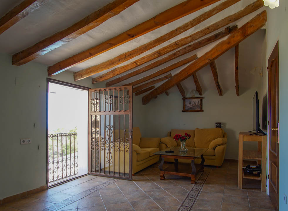 Rustic living area featuring exposed wooden ceiling beams, tiled flooring with decorative borders, and balcony access with a wrought-iron gate.