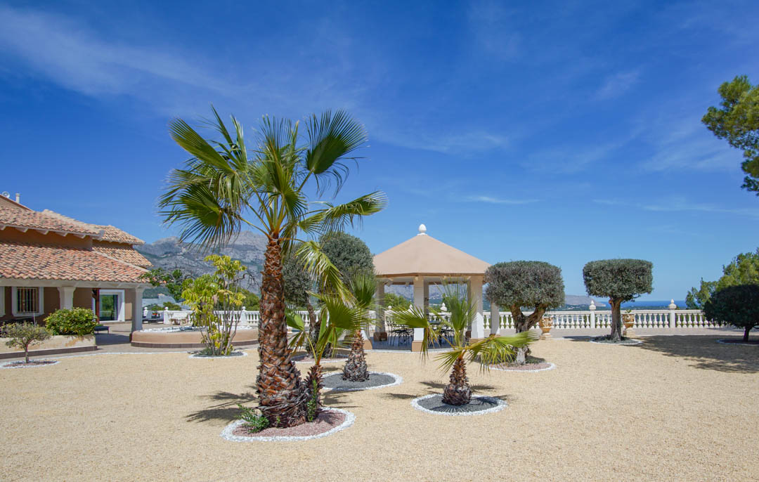 Spacious gravel courtyard featuring palm trees, manicured shrubs, and a white-columned pavilion overlooking the Mediterranean coastline and surrounding mountains.
