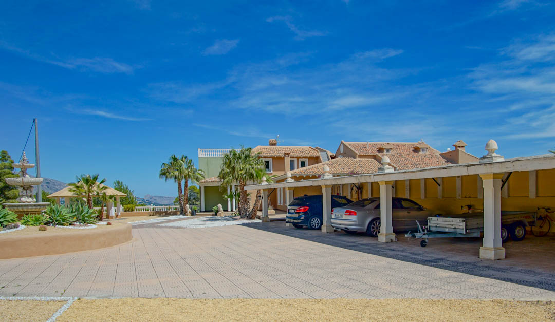 Spacious outdoor parking area featuring a covered carport for five vehicles, decorative paving, a stone fountain, and views of the main residence.
