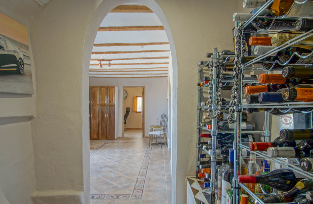 Wine storage area with chrome racks, looking through an arched passage into a room featuring traditional exposed wooden ceiling beams and tiled floors.