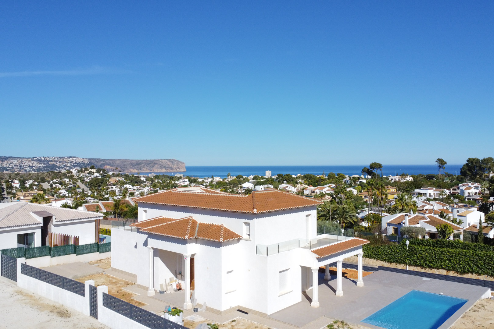 Aerial view of the white villa featuring terracotta roofing, a rectangular pool, and upper terraces with glass railings overlooking the Mediterranean Sea and coastline.