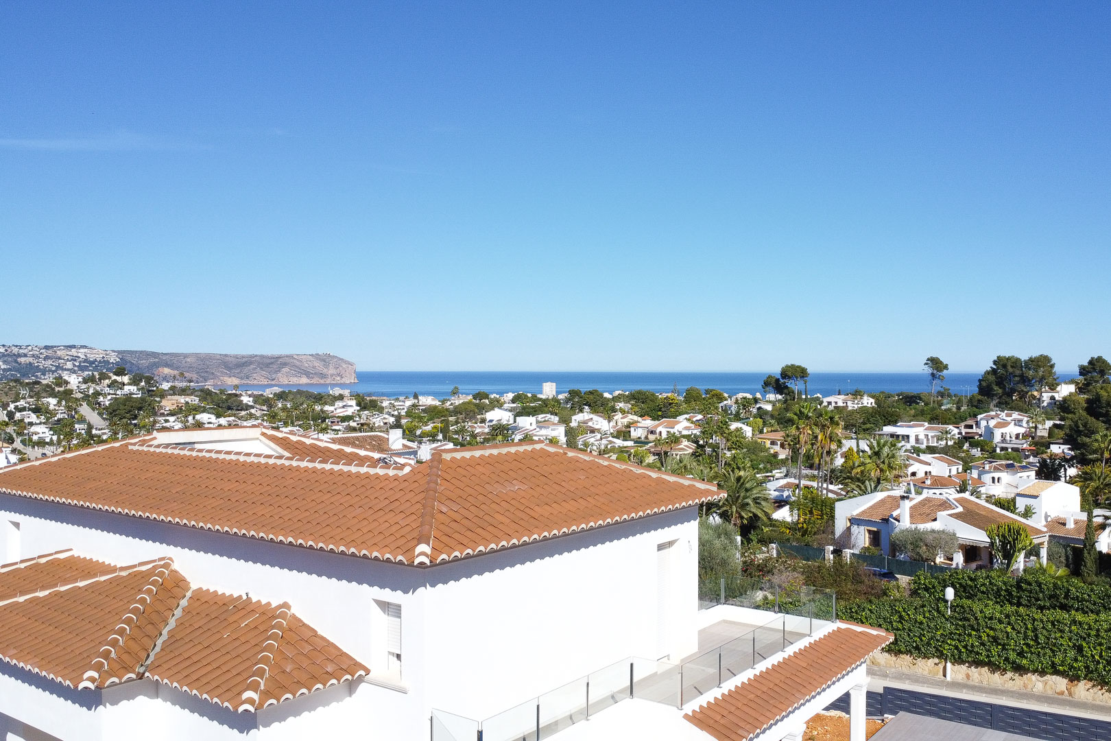 Elevated view showcasing the villa's terracotta roof, glass-railed terrace, and panoramic Mediterranean Sea views overlooking the residential landscape of Jávea.